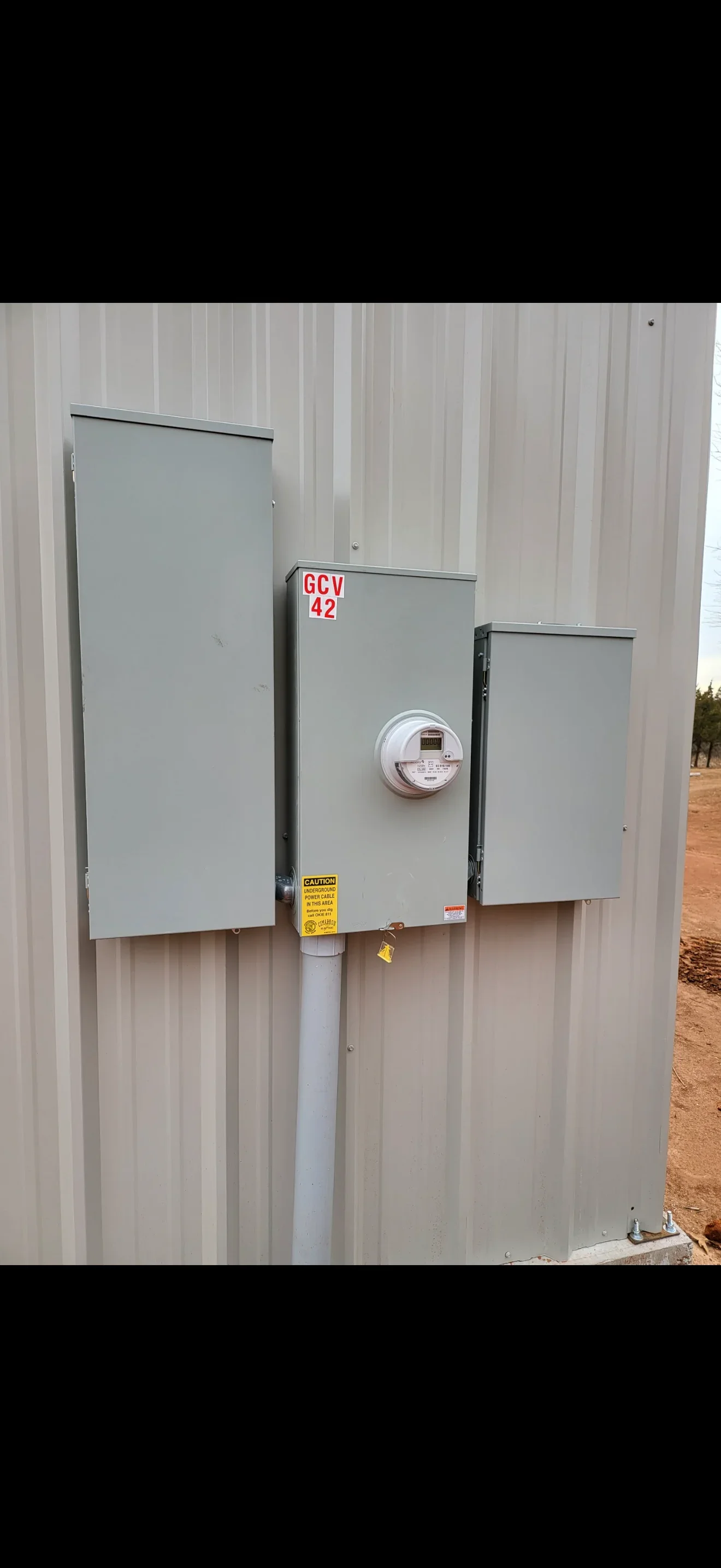 Three gray metal electrical disconnect boxes mounted on corrugated metal wall with digital meter and safety labels