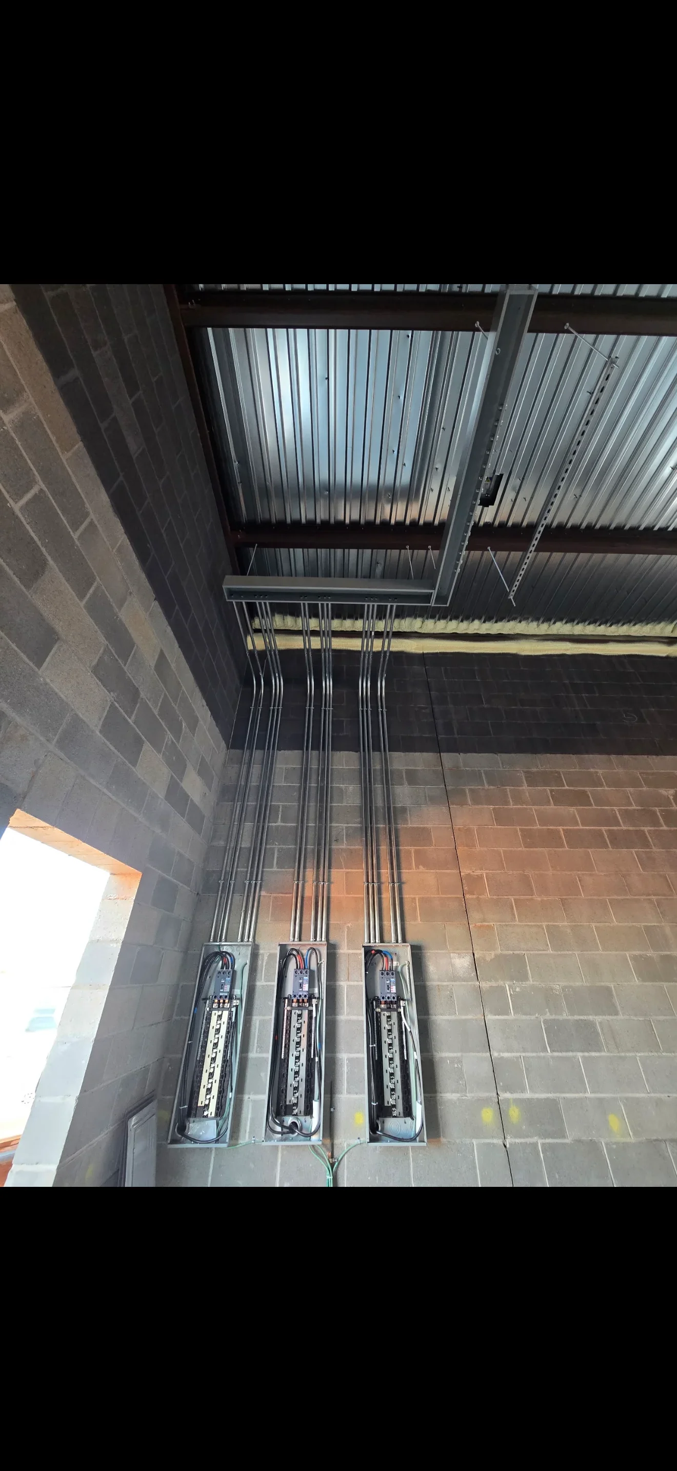 Three electrical panels with organized conduit runs on brick wall under metal roof structure